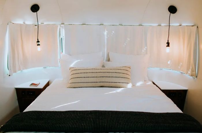 The bedroom in AutoCamp Zion's Airstreams features a queen bed dressed in a white comforter, accented by a white lumbar pillow with black geometric designs, complemented by two side tables and pendant lights hanging from the ceiling. The windows behind the bed are covered with closed curtains.