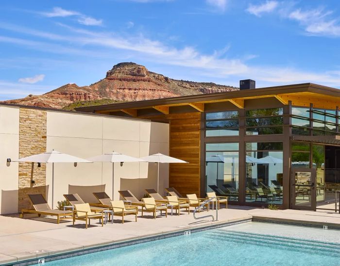 The pool area at AutoCamp Zion features a view of the pool in the foreground, adorned with six tan pool chairs and three white umbrellas to the left. In the background, the mid-century modern Clubhouse building stands, complemented by a striking red mesa behind it.