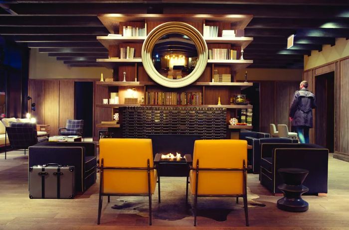 The lobby of the Thompson Chicago features black and mustard-colored chairs arranged around a cozy gas fireplace, with bookshelves positioned above.