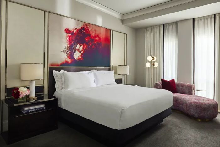 A bright guest room at the Waldorf Astoria Chicago features red artwork above the bed and a chaise longue by tall windows, creating an inviting atmosphere.