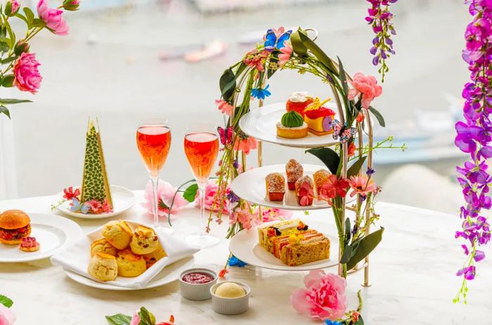 An afternoon tea display featuring a tiered tray of treats, two glasses of a pink beverage, a plate of scones, and an edible model of a skyscraper.