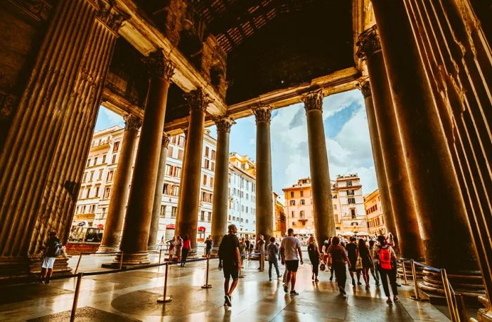 A view from inside the historic Pantheon temple in Rome, with visitors walking among the towering columns and taking in the surrounding cityscape outside.