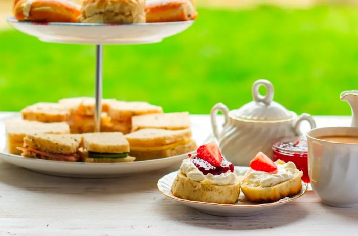 A platter of scones accompanied by clotted cream and jam, placed in front of a tray filled with tea sandwiches.