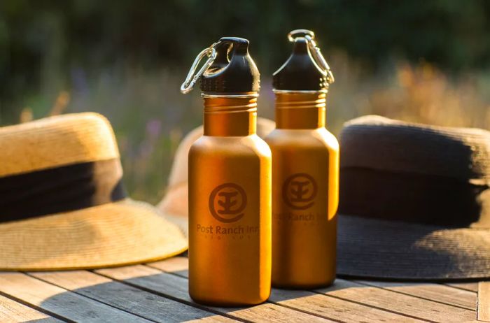 Two yellow stainless steel water bottles featuring the Post Ranch Inn logo placed in front of two hats.