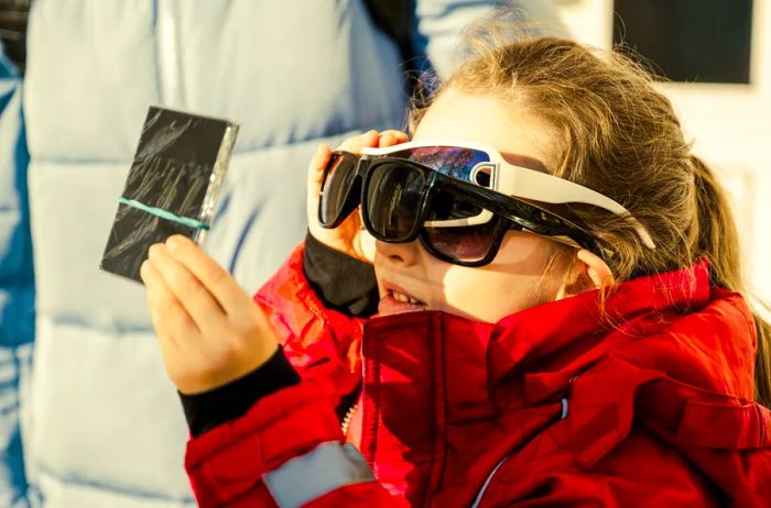 A young girl wearing multiple pairs of sunglasses along with an eclipse viewing glass to safely observe a solar eclipse.