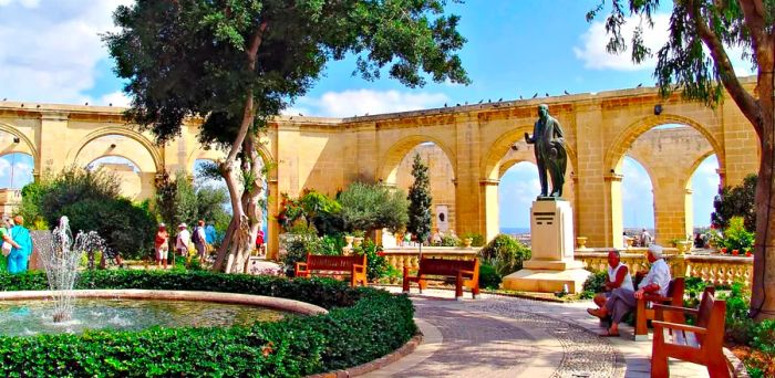 Two men relax on a bench in front of a statue, gazing at a fountain to the left in the Upper Barrakka Gardens of Valletta, Malta. Behind them, a pale yellow sandstone wall adorned with archways creates a picturesque backdrop.