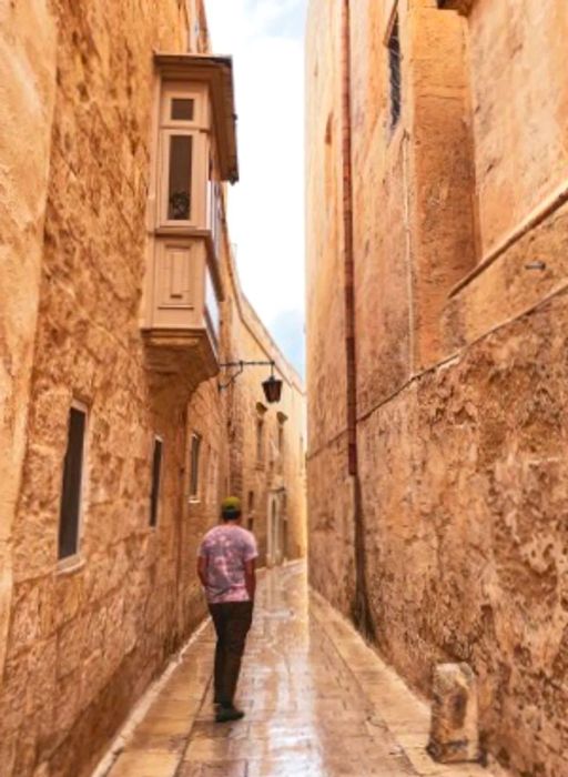 A man strolls along the stone streets of Mdina, Malta, flanked by the tall sandstone walls characteristic of the region.