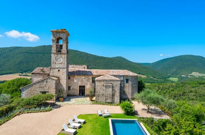 Tenuta di Murlo, a charming stone structure with a pool, set against the lush green hills of Umbria, Italy, basking in the sunshine beneath a clear blue sky.