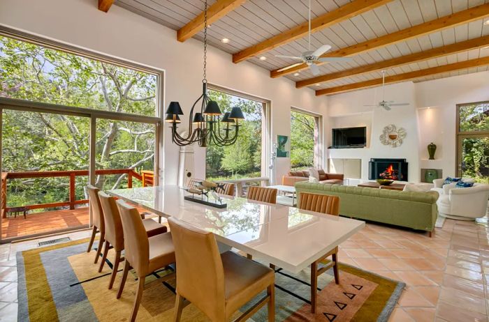 A cozy living and dining area featuring large windows, a wooden beam ceiling, a spacious dining table with chairs, and a green couch positioned in front of a kiva-style fireplace.