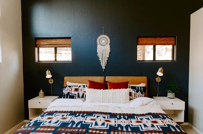 A large bed adorned with a Native American-inspired textile, positioned under a white yarn dreamcatcher, set against a navy blue wall with two small windows.