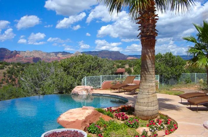 An infinity pool with a freeform design is nestled beside a small flower garden and a palm tree, set against a backdrop of red-rock mountains and cloudy blue skies.