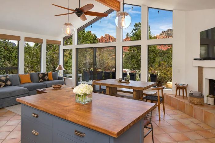 A kitchen featuring a wooden island opens up to a living area with a cozy couch and a small dining table, complemented by a double-height window that showcases views of the red rocks and vibrant desert plants.