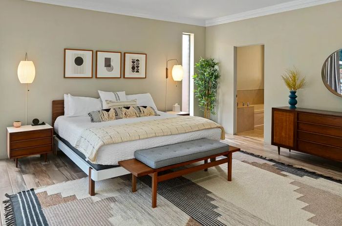 A beige bedroom featuring a midcentury wooden console and nightstand, adorned with three framed artworks on the wall, complemented by an area rug with geometric patterns.