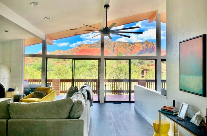 A cozy living room featuring a large sectional couch, a ceiling fan, and a spacious trapezoidal window showcasing views of the red rock mountains, lush greenery, and a blue sky dotted with white clouds.