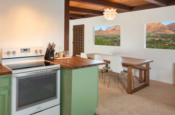 The kitchen boasts green cabinets and a white stove, with a dining area in the background featuring a long wooden table and modern white chairs, complemented by mountain views visible from two windows.