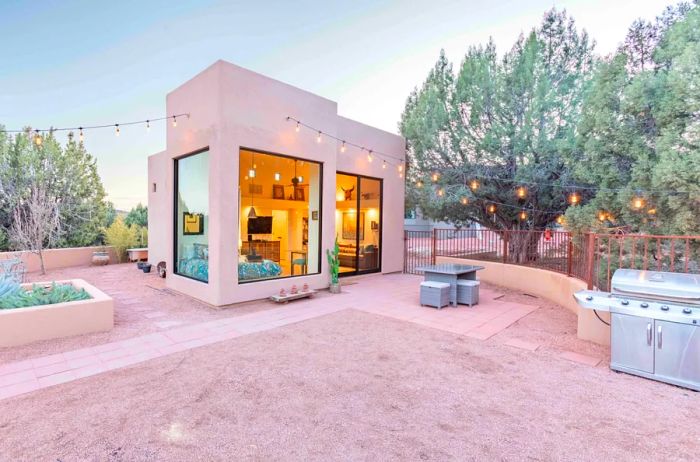 A small angular house with large windows set behind a spacious yard adorned with string lights, a small outdoor table, and a silver barbecue grill.