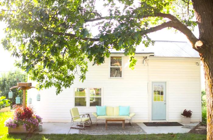 A two-story white cabin featuring a patio adorned with vintage furniture, shaded by a large tree