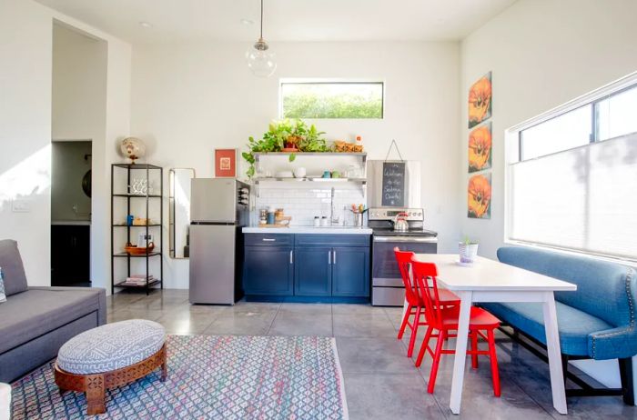 A spacious room showcasing a couch and ottoman on the left, a kitchen along the back wall, and a dining area on the right with a blue bench and bright red chairs.