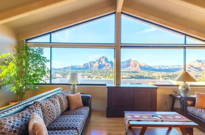 A cozy living room featuring a potted tree, a patterned sofa, a table adorned with a fabric runner, and mountain views visible through a double-height window.