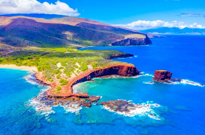 Aerial view of Lānaʻi, Hawaii showcasing Hulopo'e Bay and beach, Sweetheart Rock (Pu'u Pehe), Shark's Bay, and the mountains of Maui in the backdrop.