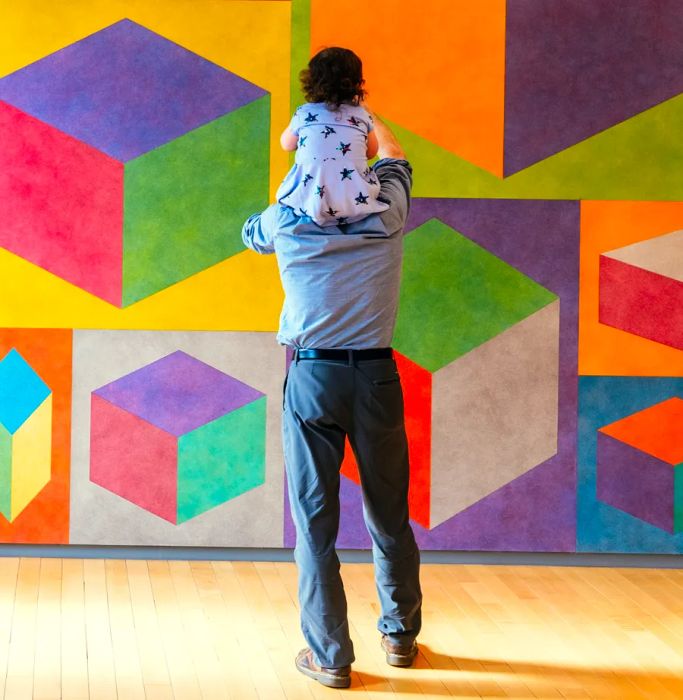 A person is seen carrying a small child on their shoulders while facing a vibrant mural of colorful cubes on a wall.