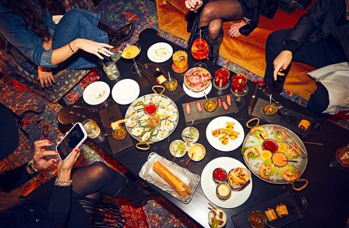 A beautifully arranged feast on a bar table, surrounded by people (visible only from the shoulders down) enjoying the spread.