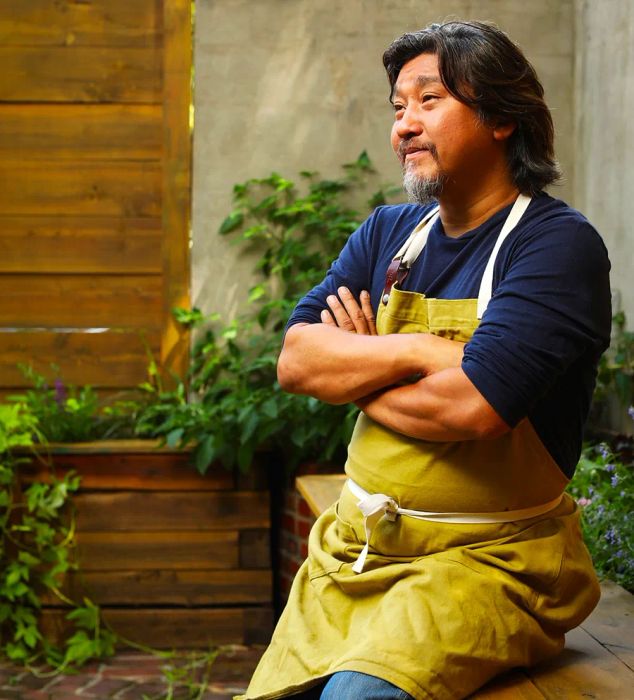 Chef Edward Lee, dressed in a yellow apron, relaxes on a bench in his backyard garden