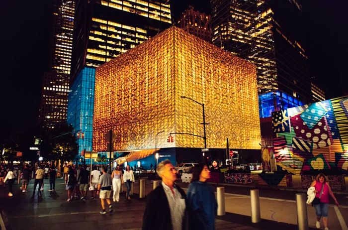 The Perelman Performing Arts Center, a cube-shaped structure, illuminates the night with a warm yellow-white glow, surrounded by buildings and bustling sidewalks.