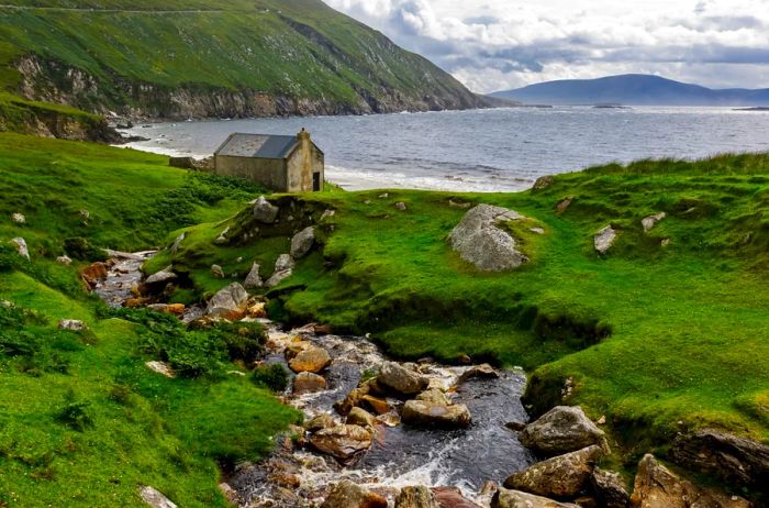Lush green hills and a rocky stream flowing down to an ancient stone house with a bay in the distance