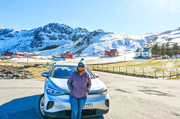 A woman stands in front of a white car, with snowy hills as the backdrop in Ulsteinvik, Norway.
