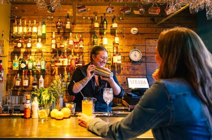 A woman vigorously shaking a cocktail mixer at the bar while another woman observes across the counter.