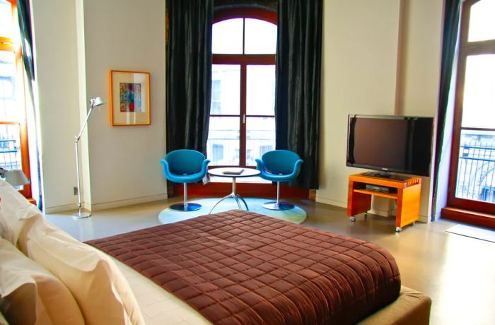 A contemporary bedroom featuring a TV on a small stand, two sleek blue chairs, expansive windows dressed with curtains, and a bed adorned with a brown grid-patterned blanket in the foreground.