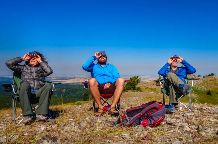Three individuals seated in camping chairs gazing up at the sky with solar eclipse viewers