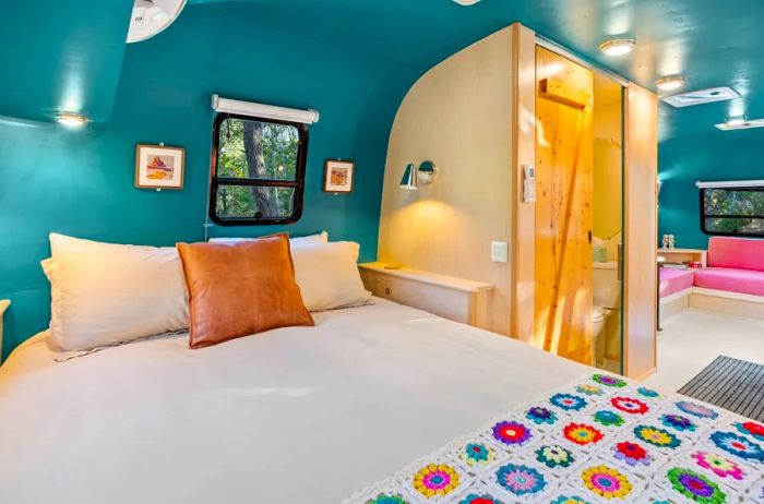 A cozy white bed adorned with a patterned crocheted blanket and a leather pillow in a trailer with green walls and a wood-enclosed bathroom