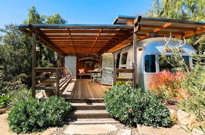 A wooden porch with a roof next to an Airstream, surrounded by lush bushes and trees