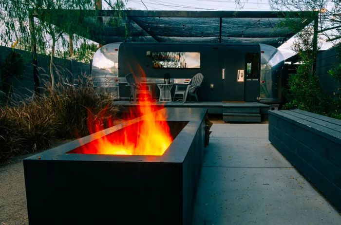 An Airstream trailer featuring a cozy porch with seating and a table, set against a modern backyard complete with a sleek black steel firepit (with flames) and a long black bench.