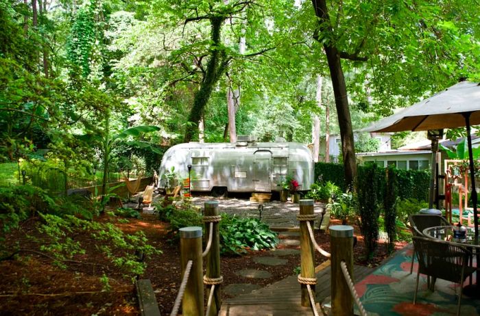 An Airstream enveloped by tropical plants in a backyard, accessible via a bridge and paver stone pathway.