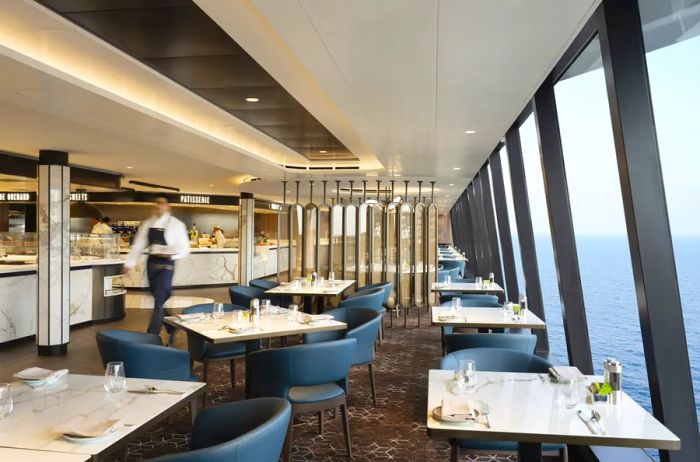 The Emporium Marketplace on the Explora I cruise ship features tables along a wall of windows with stunning sea views, while a server passes by.