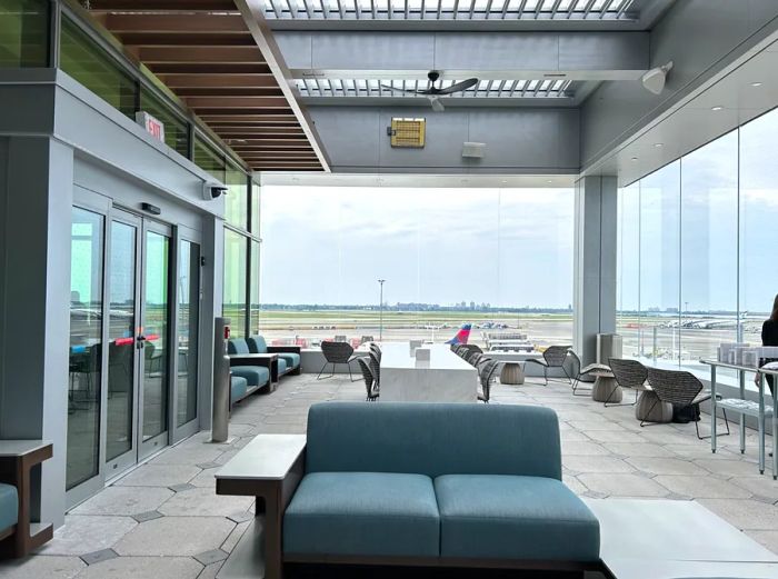 A vacant blue-gray sofa sits in the Delta Sky Club Lounge at JFK, Terminal 4, Concourse A, positioned near two walls of windows.