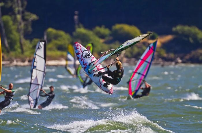 A woman gracefully soars above the Columbia River on a windsurfing board, with three other windsurfers visible in the background.