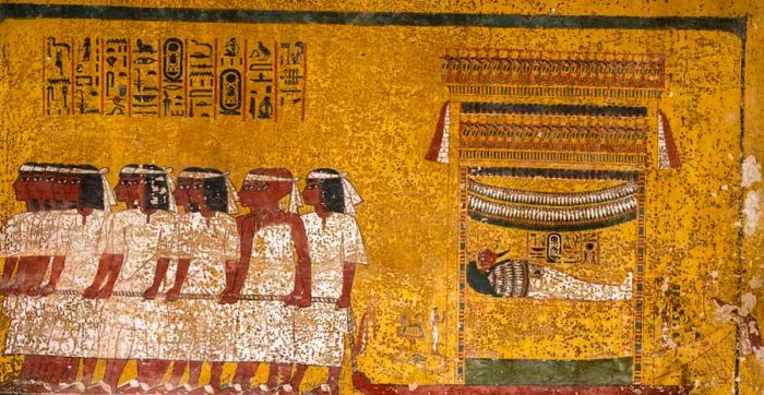 On the east wall of the tomb’s burial chamber, a scene shows Tutankhamun’s mummy lying on a shrine mounted on a sledge, drawn by twelve men in five groups. The mourners wear white bands on their heads, and the final pair, distinguished by their shaved heads and different attire, are the two viziers of Upper and Lower Egypt.