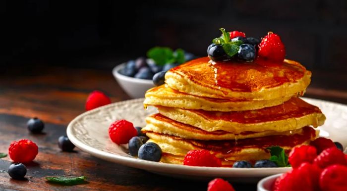 A towering stack of American pancakes topped with fresh blueberries, raspberries, and a drizzle of honey.