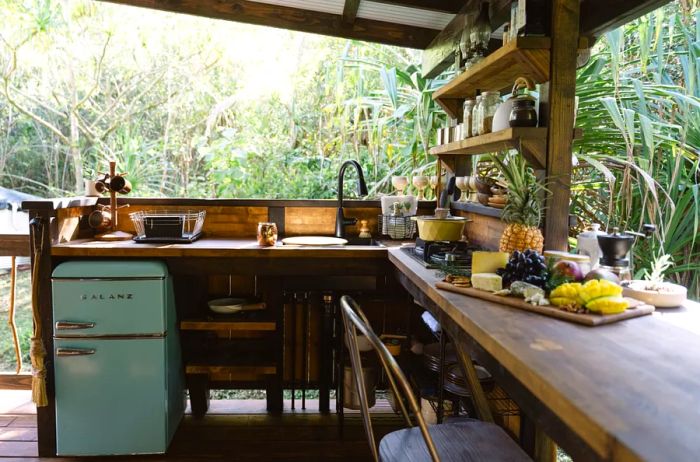 An outdoor kitchen with a long wooden table adorned with tropical fruit and a small, vintage green refrigerator, surrounded by lush tropical plants.