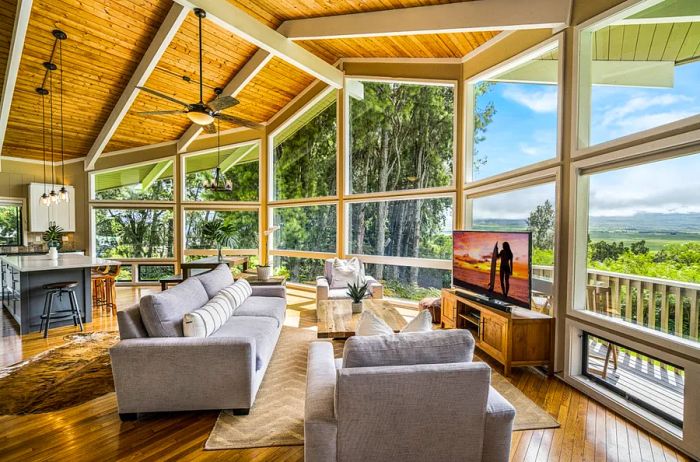 A spacious living room with wood floors and ceiling, comfortable gray couches, a large-screen TV, and floor-to-ceiling windows showcasing the lush tropical landscape outside.