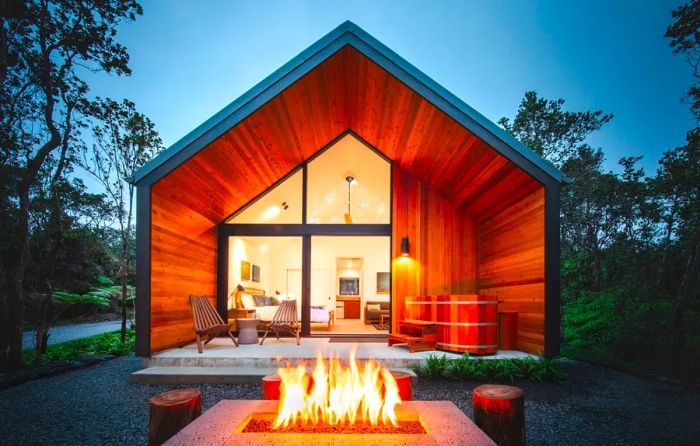 A rustic wood cabin with a sloped roof and a glass wall, featuring a glowing firepit outside at twilight.