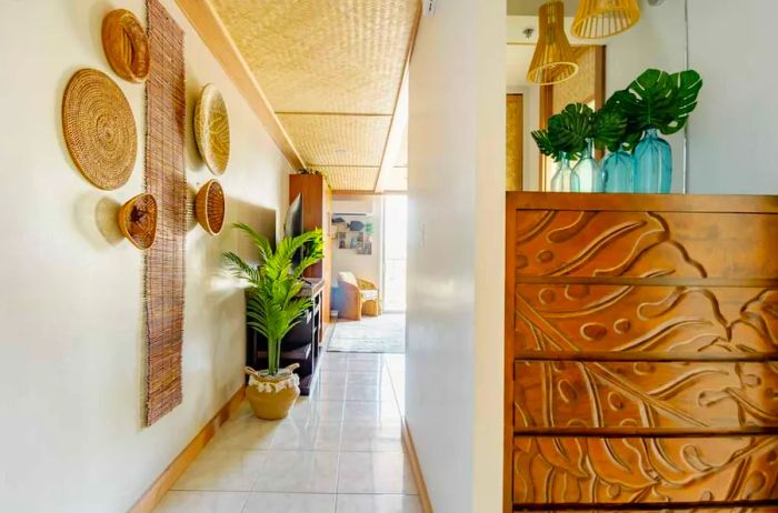 A chest of drawers carved with a monstera leaf pattern sits next to a hallway featuring a tall potted plant and baskets mounted on the wall.