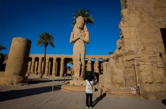 A visitor captures a photo of the iconic statue of Ramses II standing guard at the entrance to Karnak Temple.