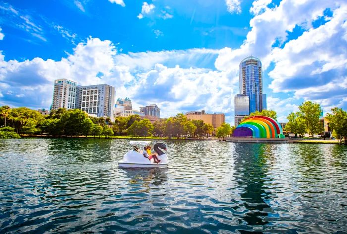 People enjoying a swan-shaped boat ride on a lake, with towering skyscrapers in the background.