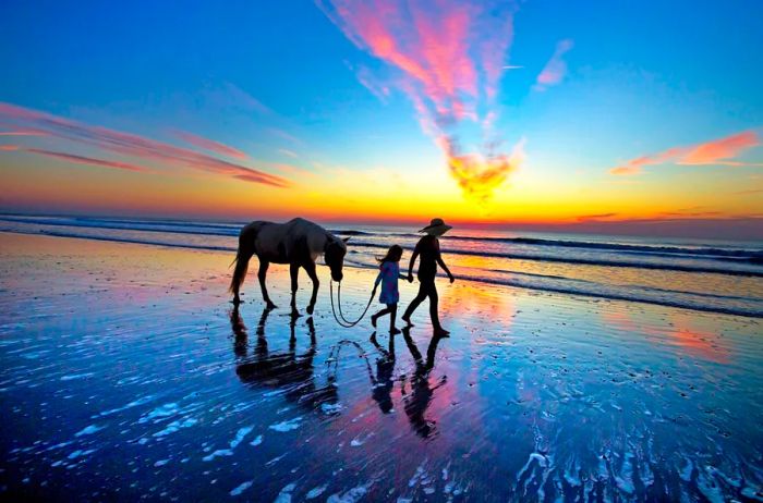 Two people stroll along the beach at sunset, walking beside a horse.