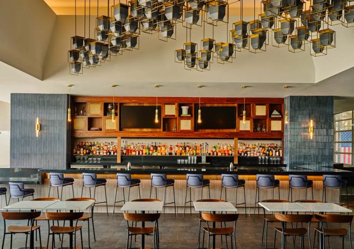 A well-stocked bar with wooden cabinetry, blue tiled accents, hanging sconces, and rows of bar stools, all set against a backdrop of empty dining tables and chairs.